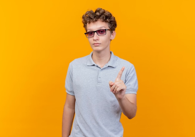 Giovane uomo con gli occhiali neri che indossa una polo grigia con il viso serio che mostra il gesto di avvertimento del dito indice in piedi sopra la parete arancione