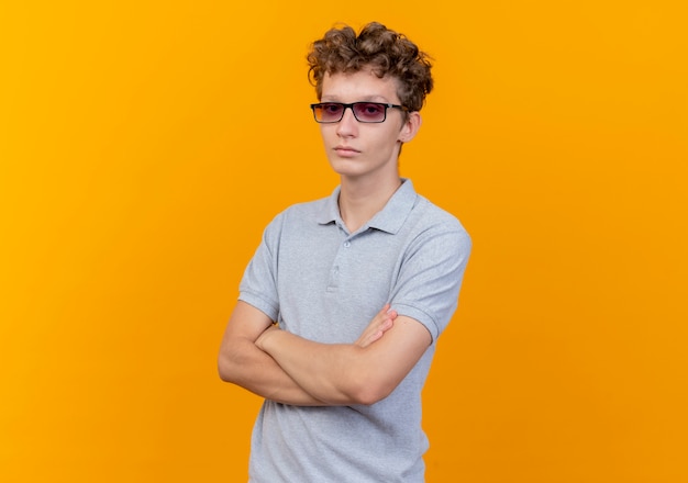 Giovane uomo con gli occhiali neri che indossa una polo grigia con il viso serio con le mani incrociate sul petto in piedi sopra la parete arancione