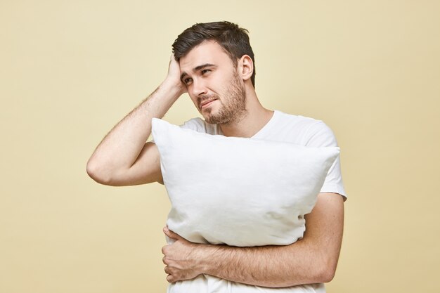 Giovane uomo con la barba lunga frustrato insoddisfatto che si sente male avendo un terribile mal di testa in posa isolato, abbracciando il cuscino, non dormendo a causa di emicrania o suoni rumorosi, avendo stressato sguardo doloroso