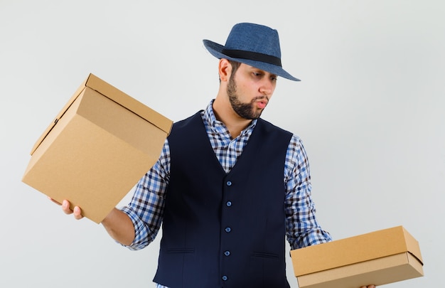 Giovane uomo con scatole di cartone in camicia, gilet, cappello e guardando curioso, vista frontale.