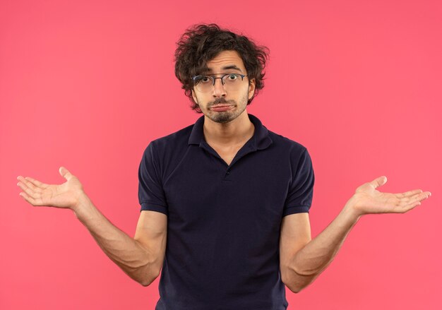 Giovane uomo confuso in camicia nera con occhiali ottici portafogli e tiene le mani aperte isolate sulla parete rosa