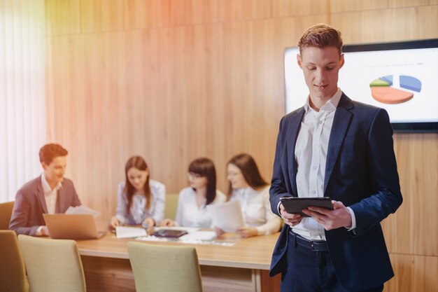 Giovane uomo d'affari alla moda che indossa una giacca e una camicia sullo sfondo di un ufficio di lavoro con persone che lavorano con un tablet