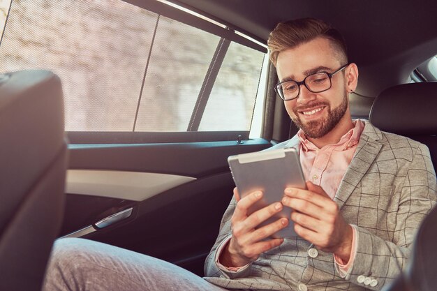 Giovane uomo d'affari alla moda di successo sorridente in un vestito grigio e una camicia rosa, usando un tablet, cavalcando su un sedile posteriore in un'auto di lusso.