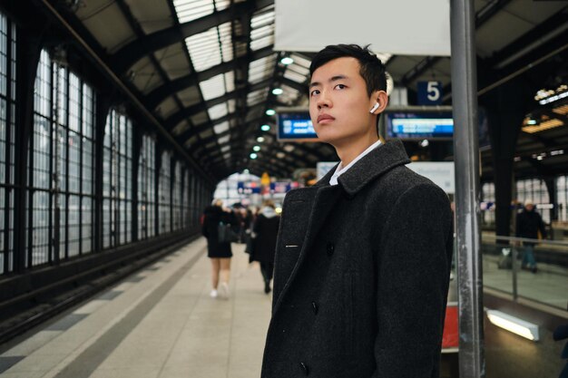 Giovane uomo d'affari asiatico alla moda in auricolari wireless in attesa del treno alla stazione della metropolitana