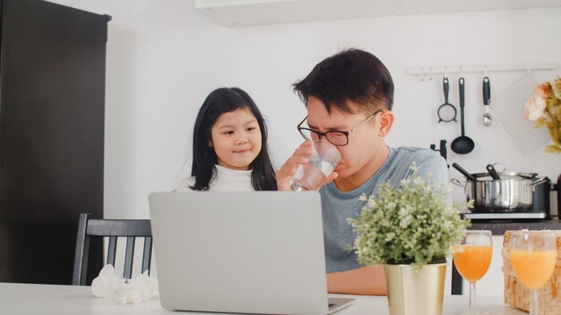 Giovane uomo d'affari asiatico serio, stress, stanco e malato mentre si lavora al computer portatile a casa. Giovane figlia che consola suo padre che lavora duramente nella cucina moderna a casa la mattina.