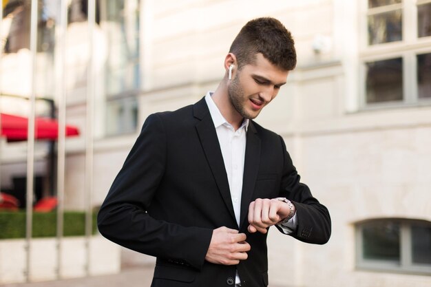 Giovane uomo d'affari attraente in abito nero e camicia bianca con auricolari wireless che guarda sognante il tempo a portata di mano mentre si trascorre il tempo all'aperto