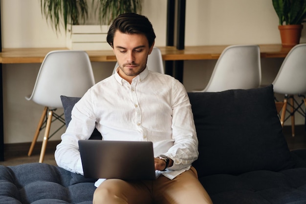 Giovane uomo d'affari attraente seduto sul divano e che lavora premurosamente sul computer portatile in uno spazio di coworking moderno