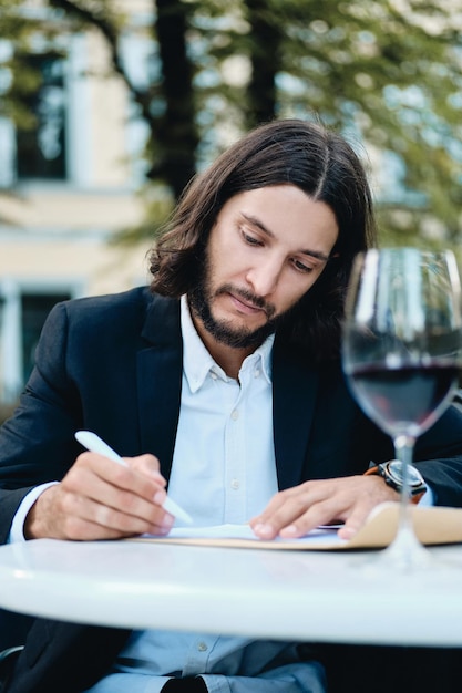 Giovane uomo d'affari barbuto attraente con bicchiere di vino rosso che lavora premurosamente nel ristorante all'aperto