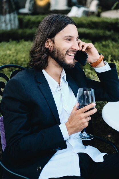 Giovane uomo d'affari barbuto bruna sorridente con un bicchiere di vino rosso felicemente parlando sul cellulare nel ristorante all'aperto
