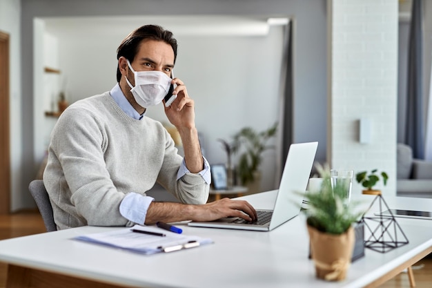 Giovane uomo d'affari che indossa una maschera protettiva mentre lavora su un laptop e parla al telefono cellulare a casa