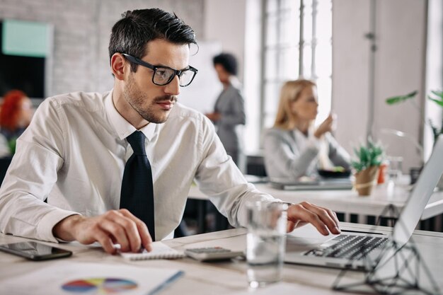 Giovane uomo d'affari che lavora su rapporti aziendali e utilizza il computer in ufficio Ci sono persone in background