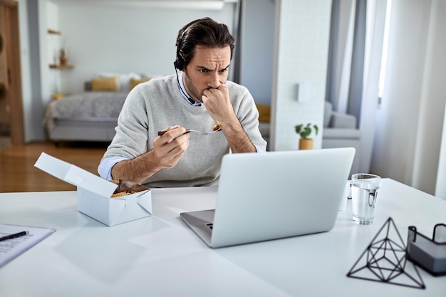 Giovane uomo d'affari che mangia mentre legge e-mail problematiche su un computer a casa