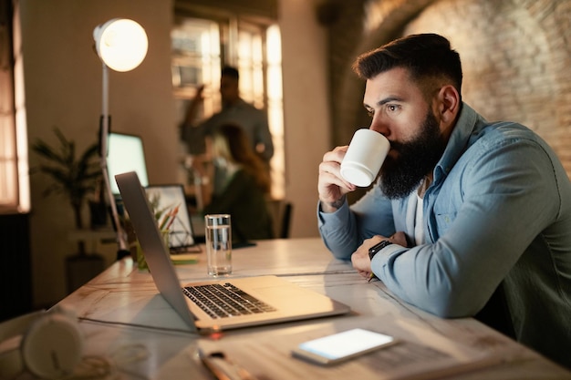 Giovane uomo d'affari che naviga in rete su un computer mentre beve un caffè la sera in ufficio