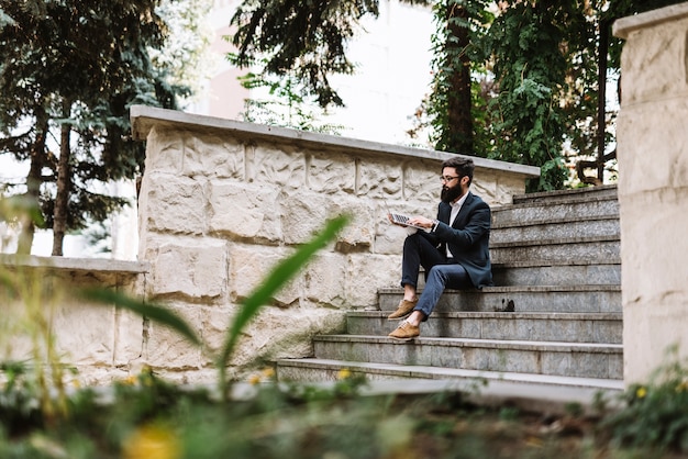 Giovane uomo d&#39;affari che si siede sulla scala per mezzo del computer portatile al parco