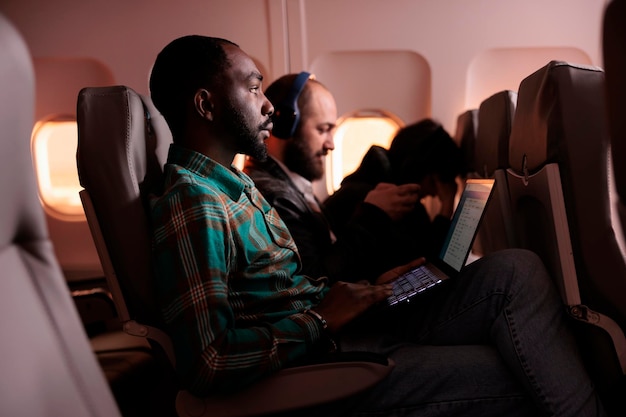 Giovane uomo d'affari che viaggia in classe economica e utilizza il laptop durante il volo, vola all'estero per una vacanza o un viaggio di lavoro. Lavorare al computer durante il tramonto, trasporto aereo.