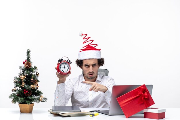 Giovane uomo d'affari con cappello di Babbo Natale e tenendo l'orologio e indicando qualcosa in ufficio su sfondo bianco