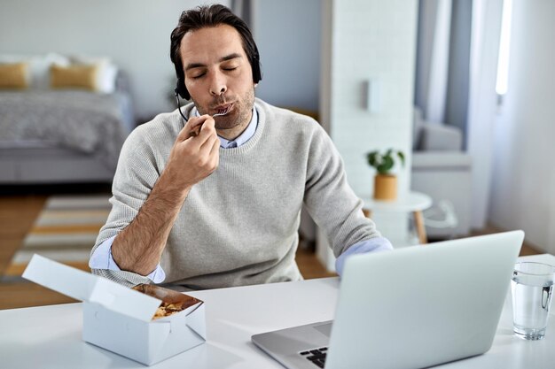 Giovane uomo d'affari con gli occhi chiusi godendosi un assaggio di cibo mentre si lavora al computer portatile a casa