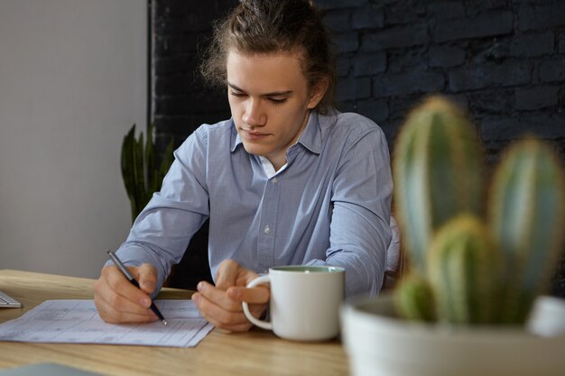 Giovane uomo d'affari concentrato laborioso in camicia blu che fa lavoro di ufficio nel suo ufficio, avendo caffè del mattino. Persone, lavoro, occupazione, professione, affari e concetto di carriera. Messa a fuoco selettiva