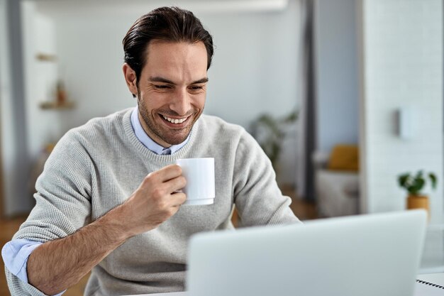 Giovane uomo d'affari felice che lavora su un computer mentre beve caffè a casa