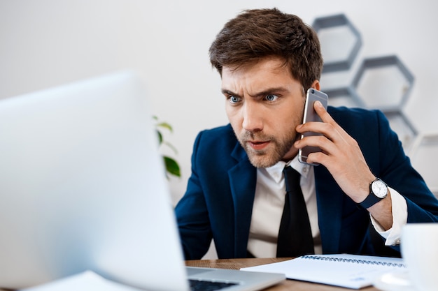 Giovane uomo d'affari infastidito che parla sul telefono, fondo dell'ufficio.