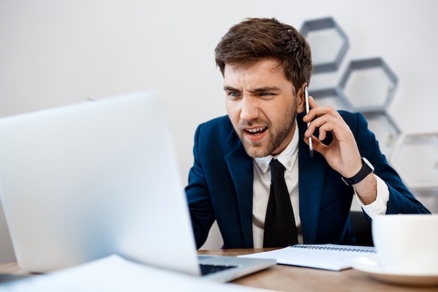 Giovane uomo d'affari infastidito che parla sul telefono, fondo dell'ufficio.