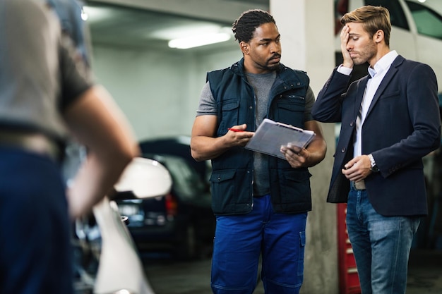 Giovane uomo d'affari preoccupato che parla con il meccanico afroamericano dei costi di riparazione dell'auto in officina