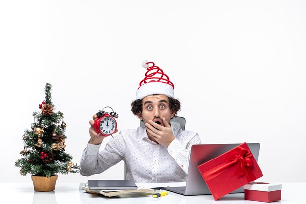 Giovane uomo d'affari scioccato eccitato con il cappello di Babbo Natale e mostrando l'orologio che lavora da solo seduto in ufficio su sfondo bianco