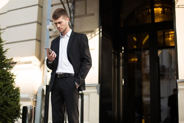 Giovane uomo d'affari serio in abito nero e camicia bianca con auricolari wireless utilizzando premurosamente il cellulare all'aperto