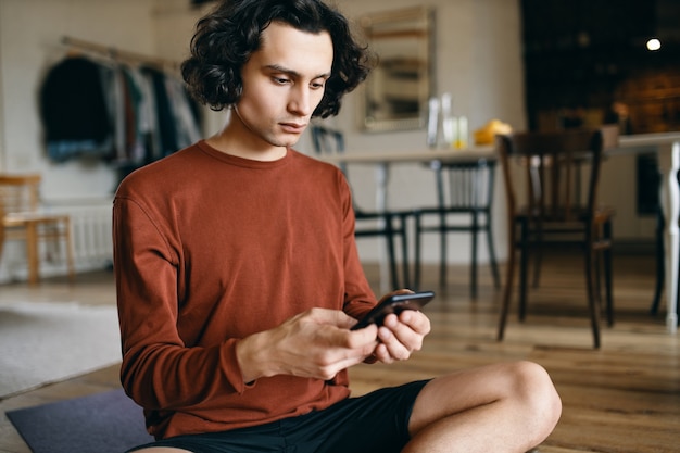 Giovane uomo d'affari serio vestito casualmente che tiene sms mobile di sms o controlla la posta elettronica mentre lavora a distanza da casa.