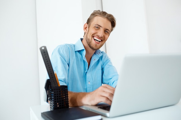 Giovane uomo d'affari sicuro allegro bello che si siede alla tavola che lavora al computer portatile con la tazza di caffè da parte. sorridente. Interno di ufficio moderno bianco