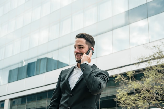 Giovane uomo d&#39;affari sorridente che sta davanti all&#39;edificio per uffici che parla sul telefono cellulare