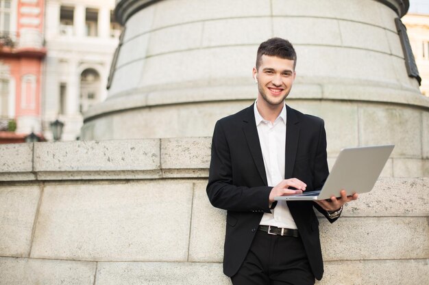 Giovane uomo d'affari sorridente nel classico abito nero e camicia bianca con auricolari wireless in piedi con il laptop in mano che guarda felicemente nella fotocamera mentre trascorre del tempo per strada