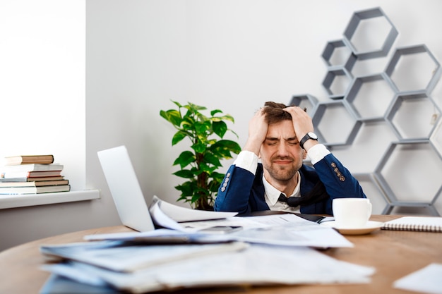 Giovane uomo d'affari turbato che si siede nel luogo di lavoro, fondo dell'ufficio.