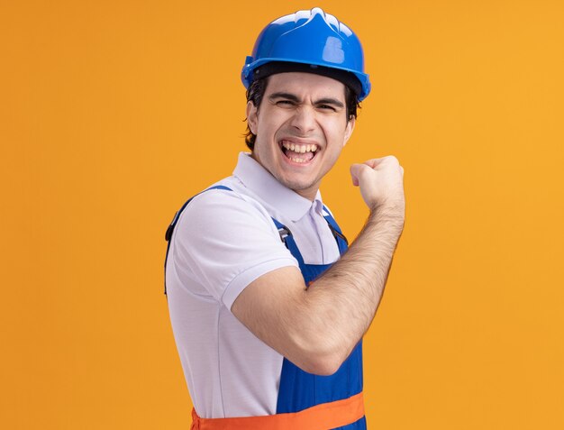 Giovane uomo del costruttore in uniforme della costruzione e casco di sicurezza che esamina il pugno di serraggio felice ed eccitato davanti che sta sopra la parete arancione