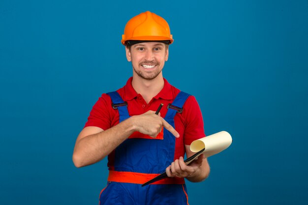 Giovane uomo del costruttore in uniforme della costruzione e casco di sicurezza che indica la lavagna per appunti in mani con il sorriso sul fronte sopra la parete blu isolata