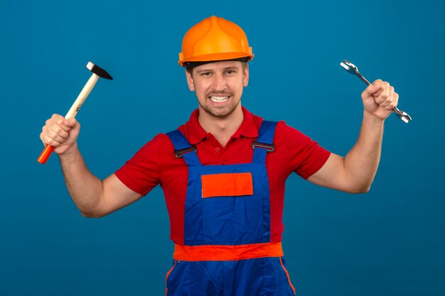 Giovane uomo del costruttore in uniforme della costruzione e casco di sicurezza che stanno con le chiavi in mani sollevate che sembrano sicure e felici sopra la parete blu isolata