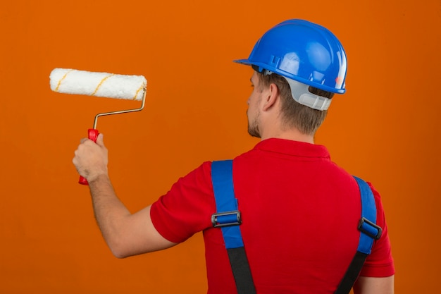 Giovane uomo del costruttore in uniforme della costruzione e casco di sicurezza che stanno indietro con il rullo di pittura sopra la parete arancio isolata