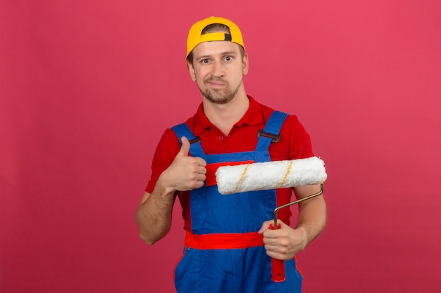 Giovane uomo del costruttore in uniforme della costruzione e rullo di pittura della tenuta del casco di sicurezza e mostrare i pollici su con l'espressione scettica sopra la parete rosa isolata