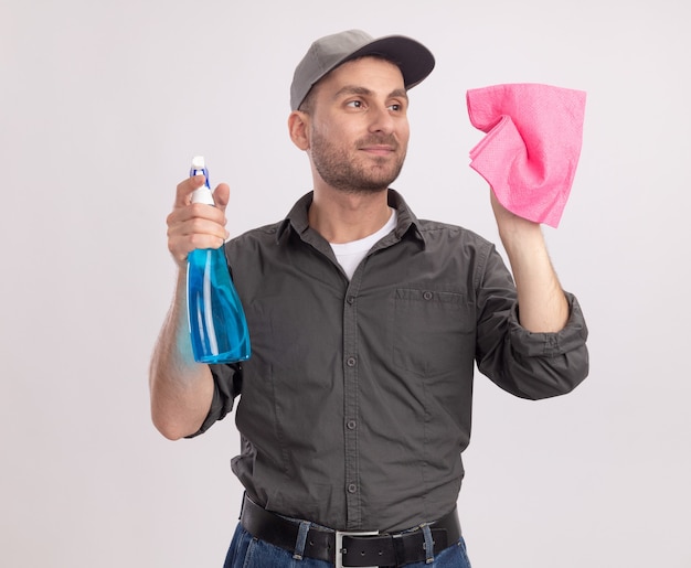 Giovane uomo delle pulizie che indossa abiti casual e berretto che tiene spray per la pulizia e straccio pronto per pulire sorridente fiducioso in piedi sopra il muro bianco