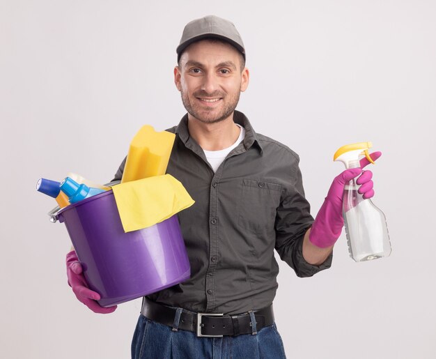 Giovane uomo delle pulizie che indossa abiti casual e berretto in guanti di gomma tenendo la bottiglia spray e secchio con strumenti per la pulizia guardando sorridente con la faccia felice in piedi sopra il muro bianco