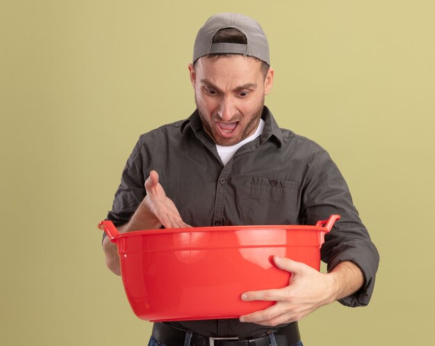 Giovane uomo delle pulizie che indossa abiti casual e berretto tenendo il bacino guardando confuso in piedi sopra la parete verde