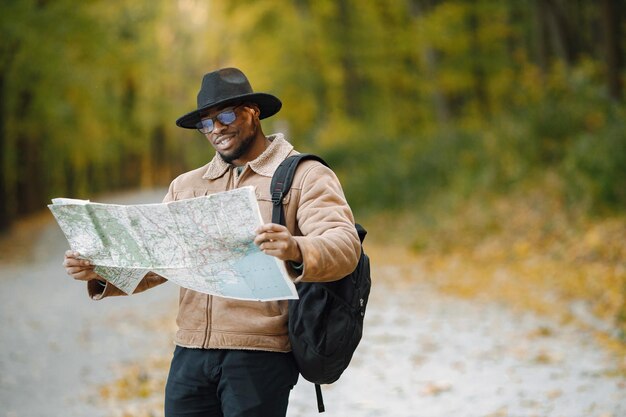 Giovane uomo di colore che fa l'autostop sulla strada e guarda la mappa. Viaggiatore maschio che si sente perso, viaggia da solo in autostop. Uomo che indossa giacca marrone, cappello nero e zaino.