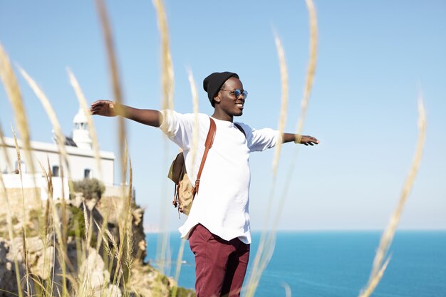 Giovane uomo di colore che indossa abiti alla moda hipster in piedi sulle rocce a picco sul mare, allargando le braccia, sentendosi spensierato e felice, sorridente, respirando aria fresca. Persone, stile di vita e viaggi