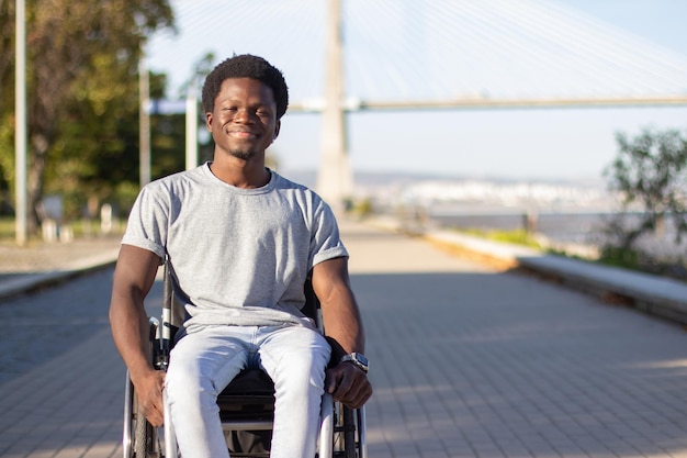 Giovane uomo di colore in sedia a rotelle che si gode una calda giornata estiva nel parco cittadino mentre si guida lungo la strada del parco. Sorridente ragazzo afroamericano con disabilità che trascorre del tempo fuori. Disabilità, concetto di stile di vita.