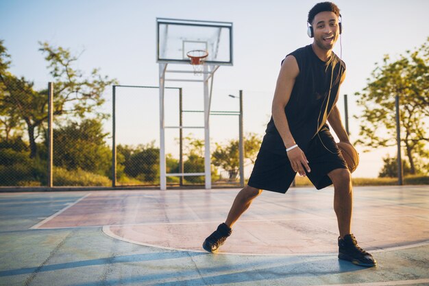 Giovane uomo di colore sorridente felice che fa sport, gioca a basket all'alba, ascolta la musica in cuffia, stile di vita attivo, mattina d'estate