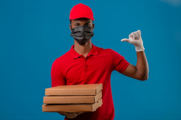 Giovane uomo di consegna afroamericano che indossa la polo rossa e il cappuccio in maschera protettiva e guanti in piedi con la pila di scatole per pizza che punta con il dito a se stesso su blu isolato