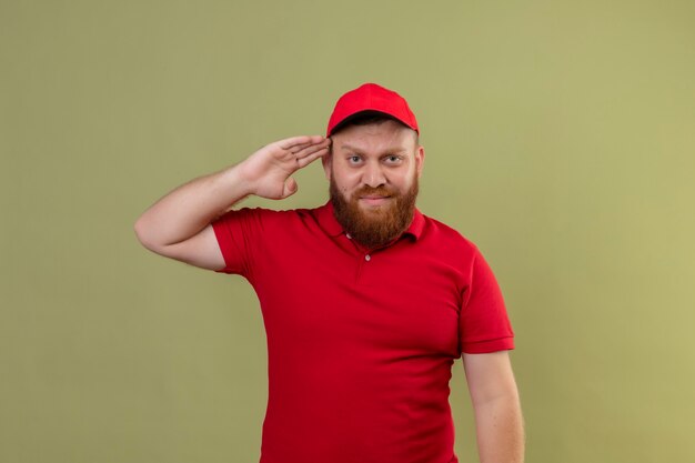 Giovane uomo di consegna barbuto in uniforme rossa e berretto che sembra salutare fiducioso