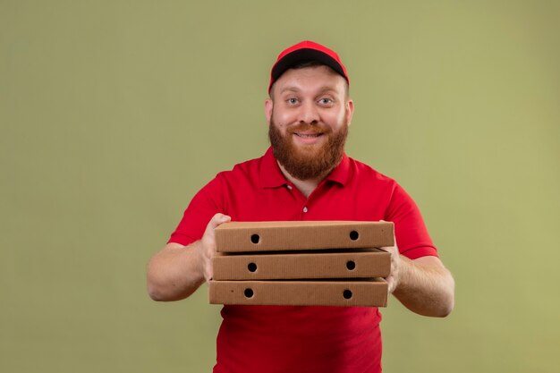 Giovane uomo di consegna barbuto in uniforme rossa e berretto hodling pila di scatole per pizza guardando la telecamera sorridente amichevole