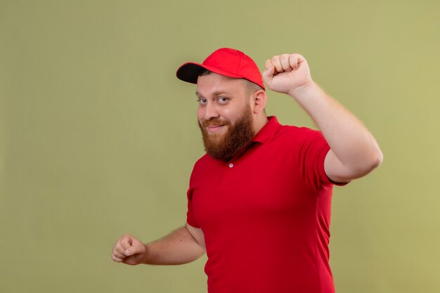 Giovane uomo di consegna barbuto in uniforme rossa e berretto sorridente pugno di sollevamento amichevole come un vincitore, felice e positivo