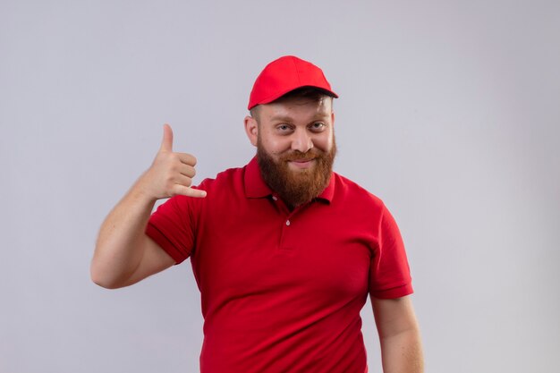 Giovane uomo di consegna barbuto in uniforme rossa e cappello sorridente facendomi cal gesto con la mano